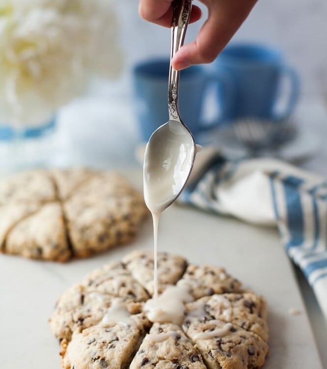 Hand holding a spoon of frosting over chocolate chip toffee scones on a white counter