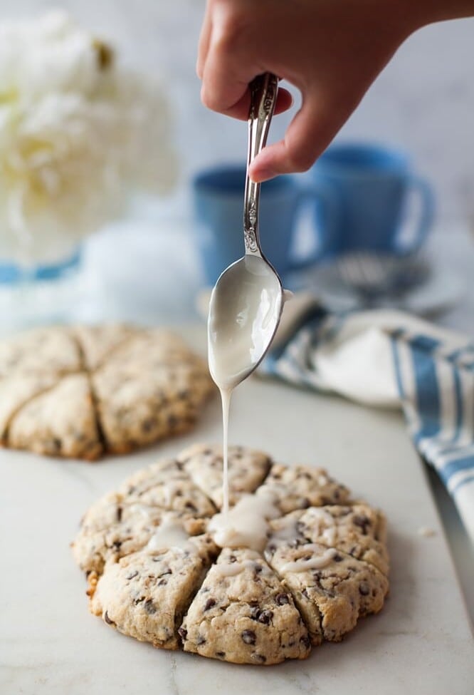 Hand holding a spoon pouring icing over a batch of scones on a marble counter top