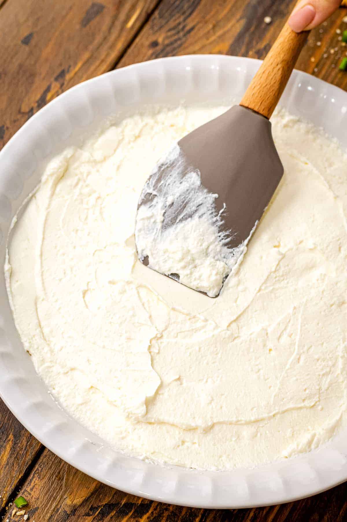 White pie plate with spatula spreading cream cheese on the bottom.