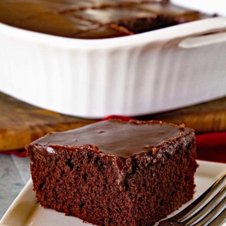 Piece of homemade chocolate cake with chocolate frosting on a white plate with a fork in front of a white baking dish containing the rest of the cake