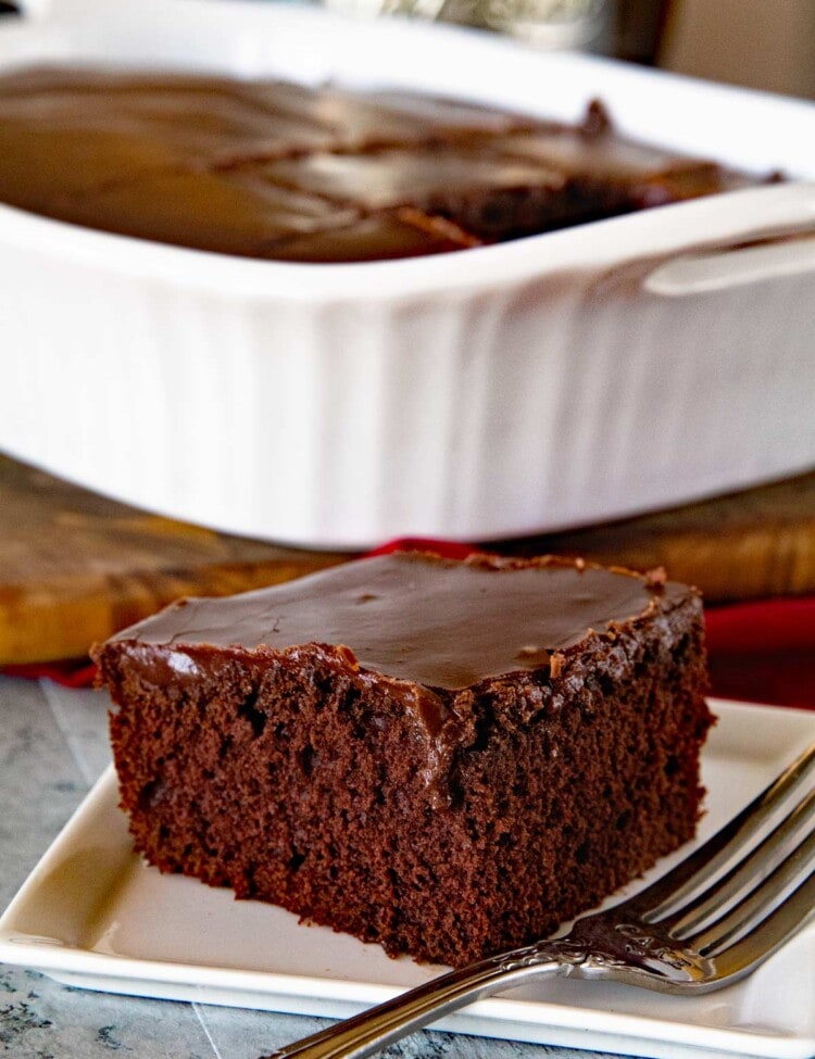A slice of chocolate cake with chocolate frosting on a square white plate with a fork