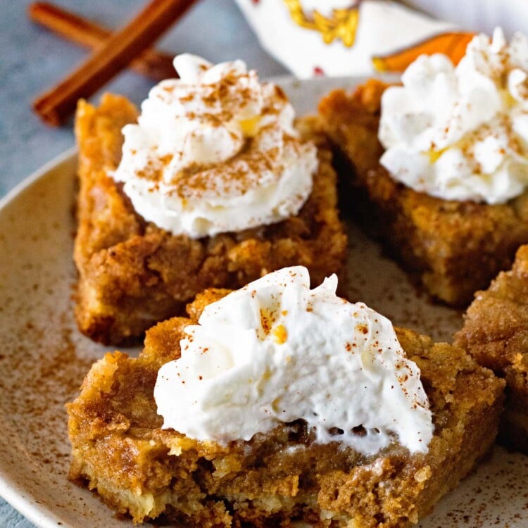 Four pumpkin pie bars on a plate topped with whipped cream and cinnamon with the front bar having a bite out of it