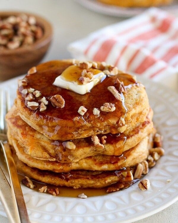 Stack of sweet potato pancakes with butter, syrup, and chopped pecans on a white plate with a fork