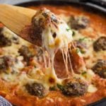 Wooden spatula lifting a meatball and cheese out of a cast iron skillet of cheesy Italian meatballs and pasta