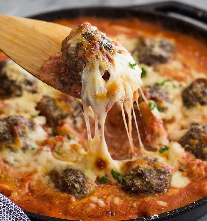 A wooden spoon lifting a cheesy meatball out of a skillet of meatballs and pasta