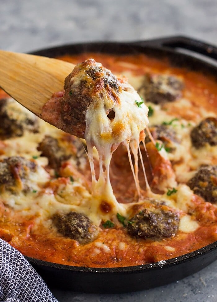 A wooden spoon lifting a cheesy meatball out of a skillet of meatballs and pasta