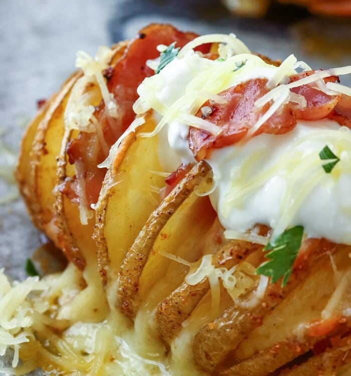 Loaded Hasselback Potato topped with sour cream and shredded cheese on a baking sheet lined with parchment paper