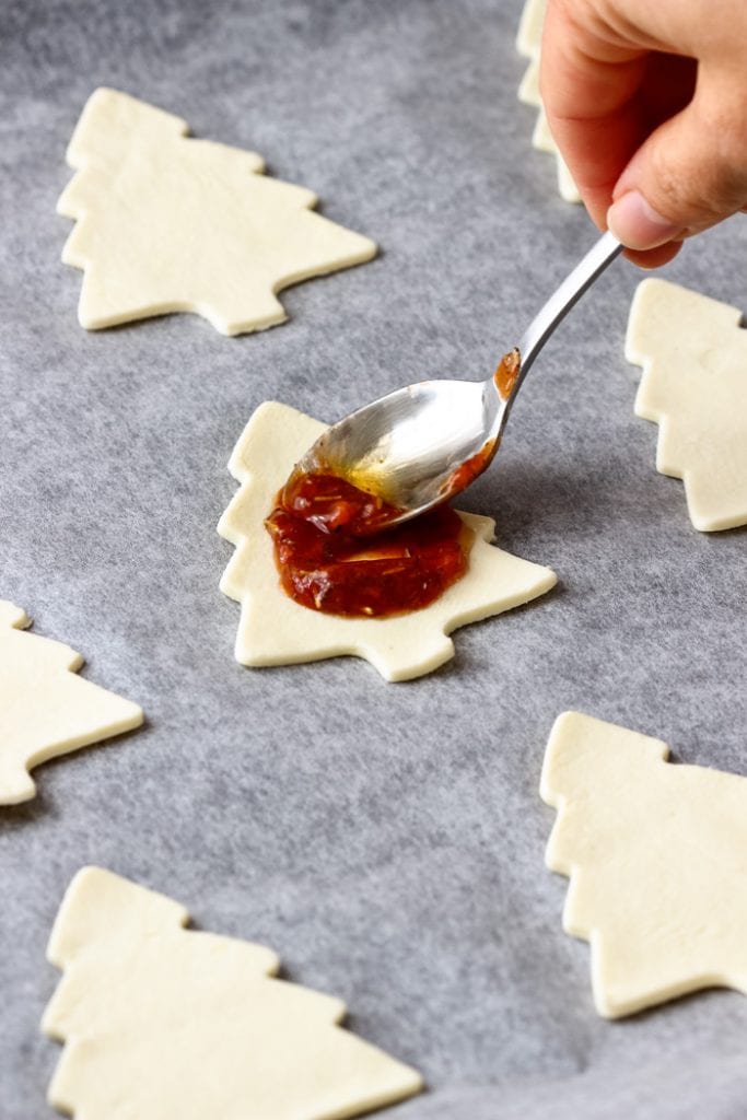 Christmas tree shaped pieces of dough being spread with pizza sauce from a spoon