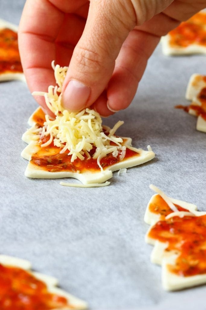 Christmas Tree Mini Pizzas with sauce on them being sprinkled with shredded cheese on a sheet pan