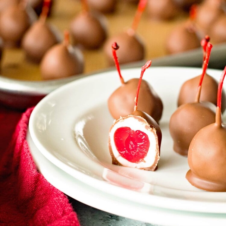 Homemade chocolate covered cherries on a white plate with one of them cut in half to show the cherry in the center