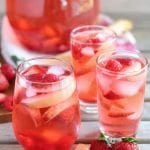 Three glasses and a pitcher of peach rose sangria on a wood table with slices of peach and strawberries
