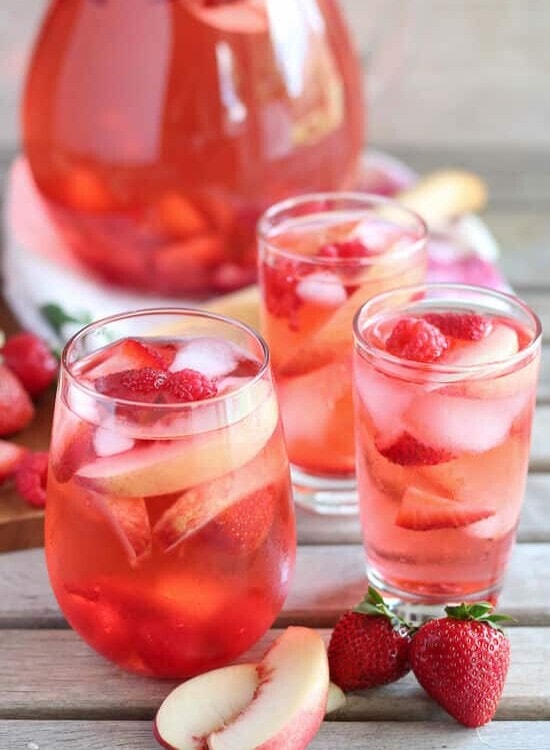 Three glasses and a pitcher of peach rose sangria on a wood table with slices of peach and strawberries