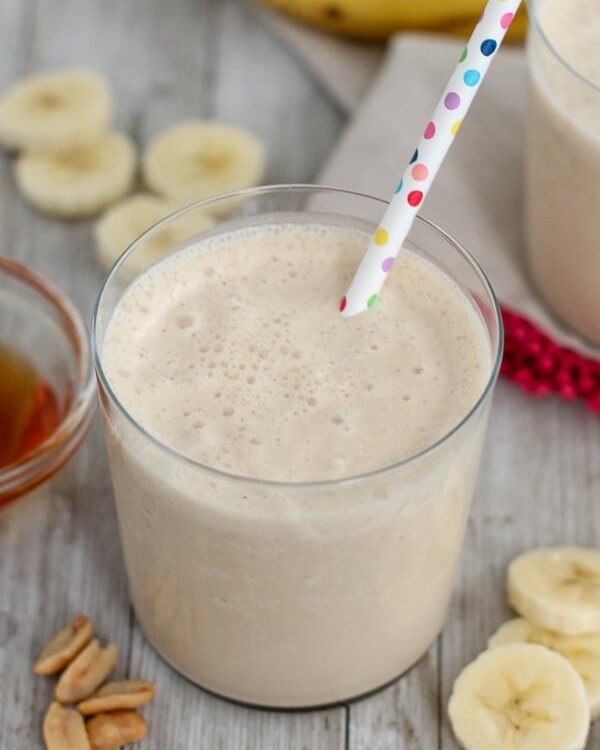 A glass of peanut butter banana smoothie with a polka dot straw in it next to slices of banana, peanuts, and a bowl of honey