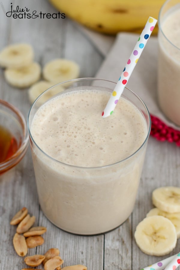 A glass of peanut butter banana smoothie with a polka dot straw in it next to slices of banana, peanuts, and a bowl of honey