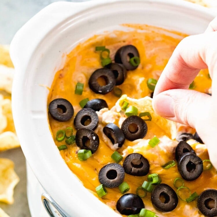 A hand dipping a chip into a white slow cooker of chicken enchilada dip topped with black olives