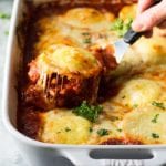 Hand holding a spatula with a ravioli on it over a white casserole dish of three cheese ravioli lasagna