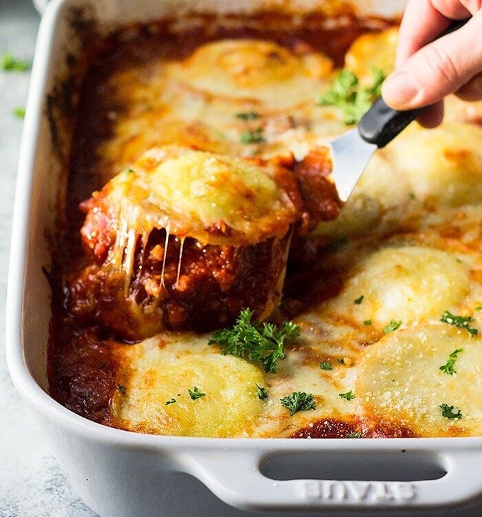 Hand holding a spatula with a ravioli on it over a white casserole dish of three cheese ravioli lasagna