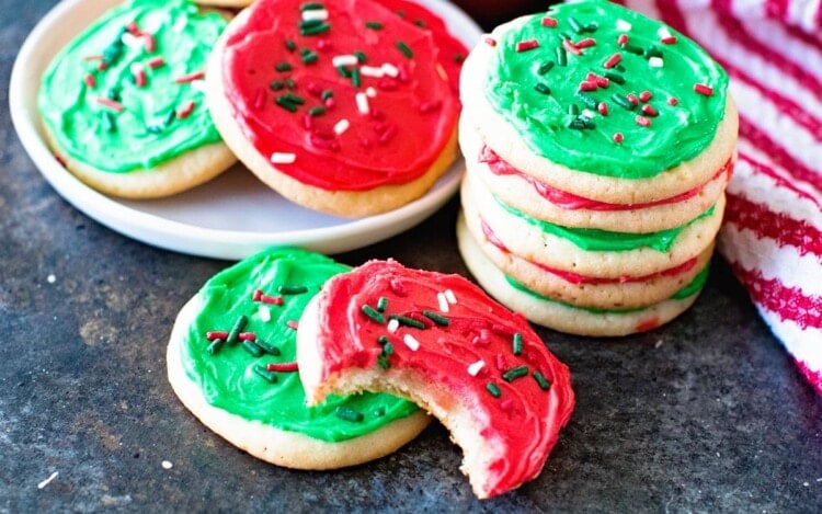 A stack of sugar cookies with red and green frosting next to two more cookies on the counter one with a bite out of it and a plate of sugar cookies with red and green frosting
