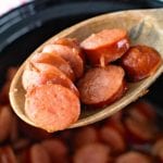 Wooden spoon with glazed kielbasa bites on it being held over a crock pot