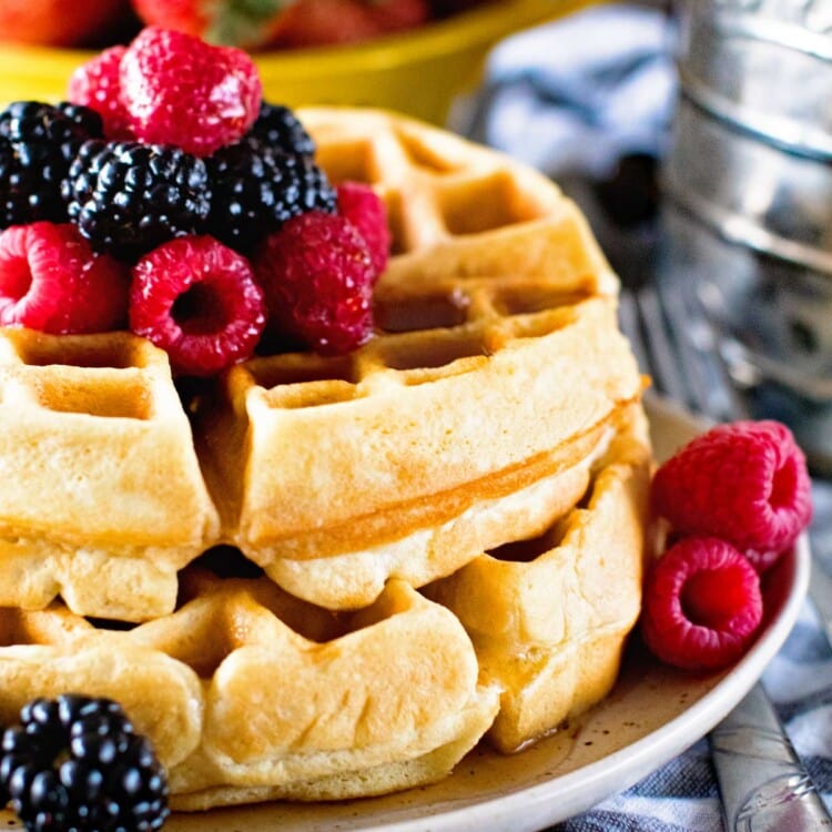 Two homemade waffles stacked on a cream plate with berries and syrup on top next to a bowl of strawberries