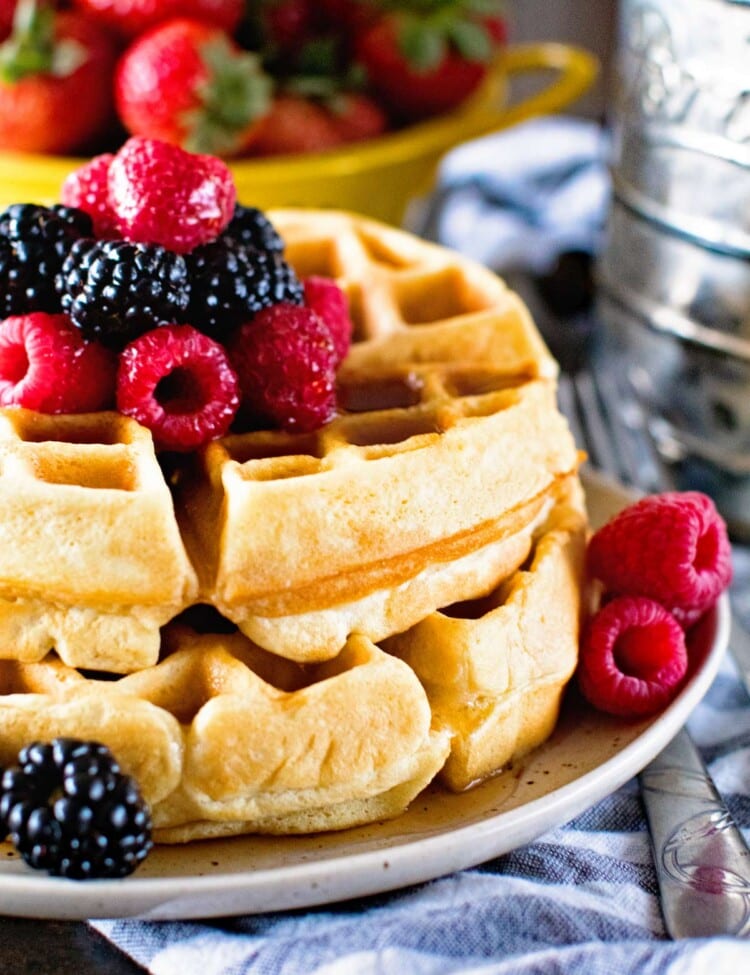 Two homemade waffles stacked on a cream plate with berries and syrup on top next to a bowl of strawberries