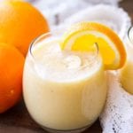 Glass of fresh orange smoothie with banana slices in it and an orange slice on the rim sitting on a wooden table with a white cloth and two oranges