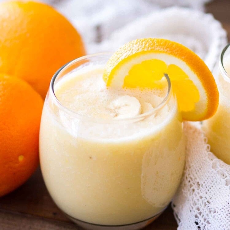 A glass of fresh orange smoothie with an orange slice on the rim next to two oranges