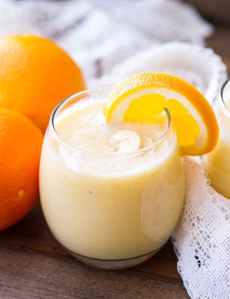 A glass of fresh orange smoothie with an orange slice on the rim next to two oranges