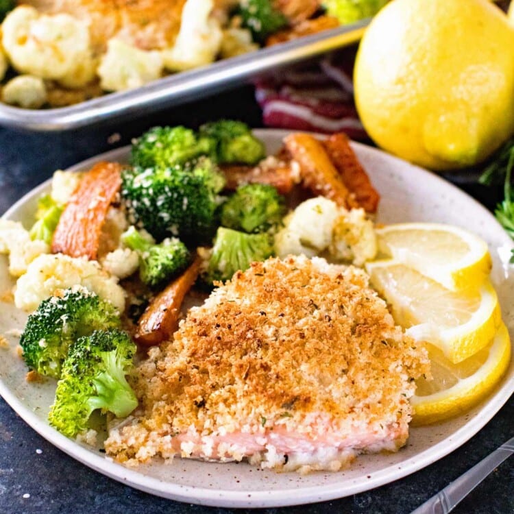 Breaded salmon, broccoli, cauliflower, and lemon slices on a plate in front of a sheet pan of salmon