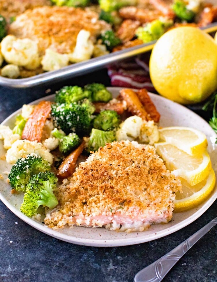 Breaded salmon, broccoli, cauliflower, and lemon slices on a plate in front of a sheet pan of salmon