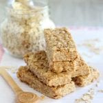 A pyramid of no bake peanut butter honey granola bars on a white plastic cutting board along with oats, a glass jar of oats, and a small wood spoon full of peanut butter