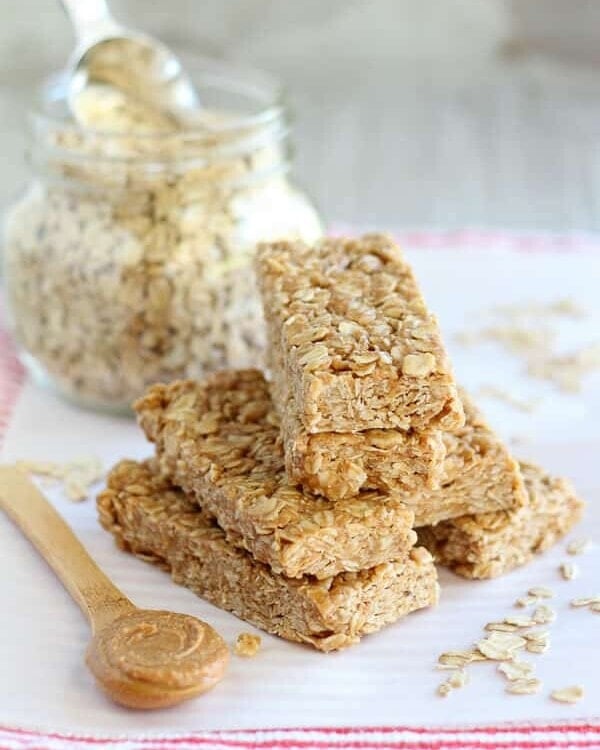 A pyramid of no bake peanut butter honey granola bars on a white plastic cutting board along with oats, a glass jar of oats, and a small wood spoon full of peanut butter