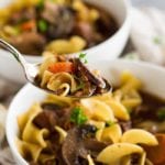 Two white bowls of slow cooker beef and noodle soup with a spoon of soup being held over one