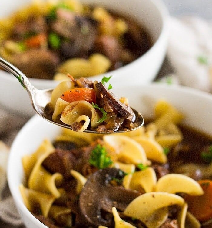 Two white bowls of slow cooker beef and noodle soup with a spoon of soup being held over one