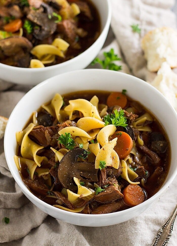 slow cooker beef and noodle soup in two white bowls on a kitchen towel
