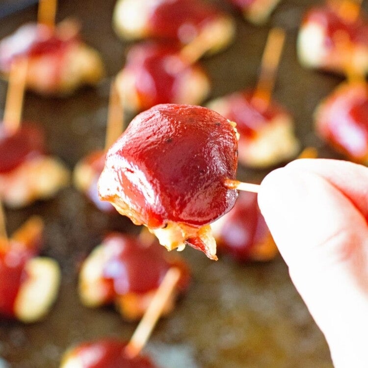 Hand holding a toothpick with a bbq bacon chicken bite on it over a pan of bbq bacon chicken bites