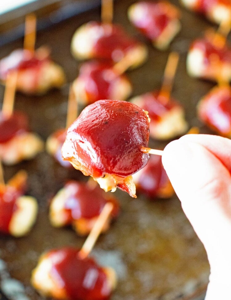 Hand holding a toothpick with a bbq bacon chicken bite on it over a pan of bbq bacon chicken bites