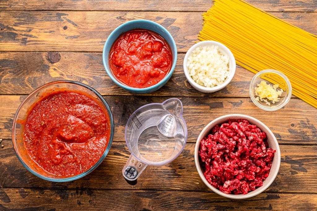 Overhead photo of ingredients in bowls including spaghetti sauce, diced tomatoes, water, garlic, ground beef, spaghetti noodles etc.