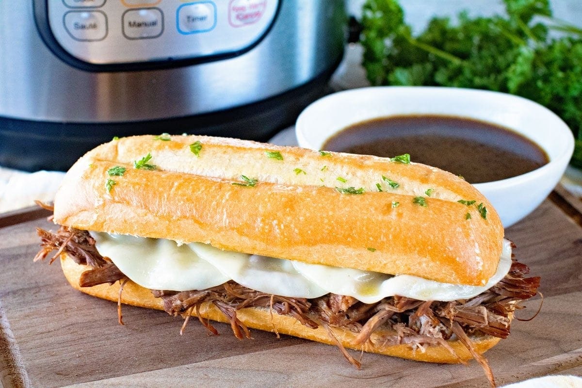 French Dip Sandwich next to a white bowl of sauce on a wood cutting board in front of a pressure cooker