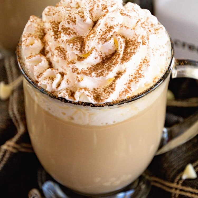 A clear glass mug of white chocolate latter topped with whipped cream and cinnamon