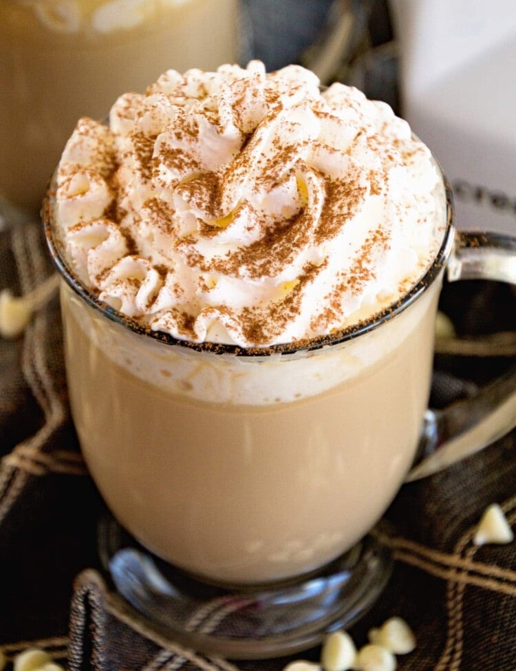A clear glass mug of white chocolate latter topped with whipped cream and cinnamon