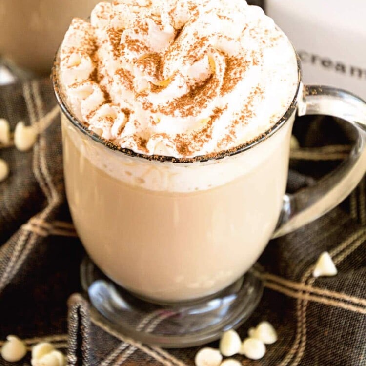 A clear glass mug of white chocolate latte with whipped cream and cinnamon on top sitting on a cloth napkin with white chocolate chips