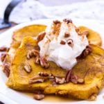 two slices of baked pumpkin french toast with syrup, pecans, and whipped cram on top on a white plate