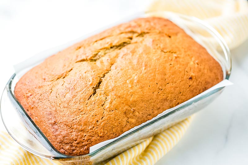 Glass loaf pan with a baked loaf of banana bread in it on top of yellow and white striped napkin.