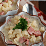 Two bowls of crock pot cream cheese ham and potato soup on a wood board with a spoon