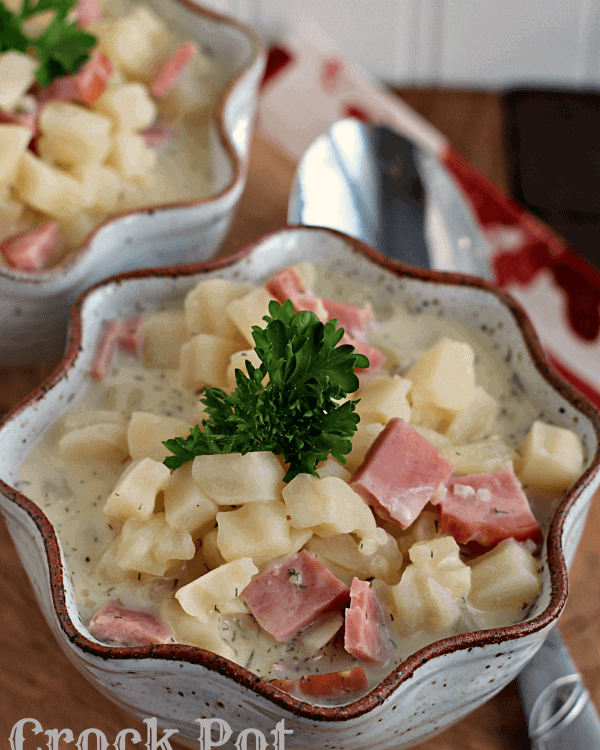 Two bowls of crock pot cream cheese ham and potato soup on a wood board with a spoon
