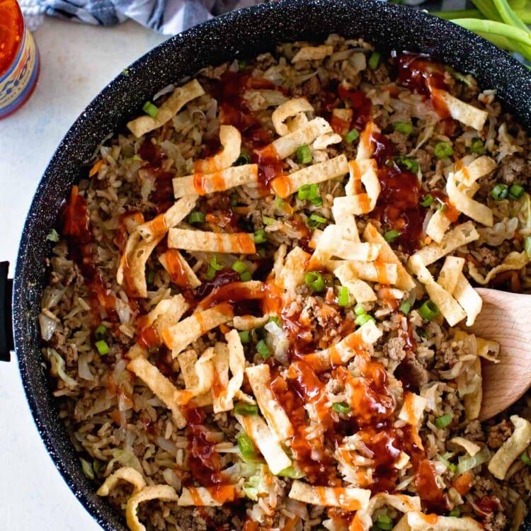 Overhead image of one pan egg roll in a bowl being stirred with a wood spoon next to a bottle of sauce, a bowl of crunchy strips, an onion, and some green onions
