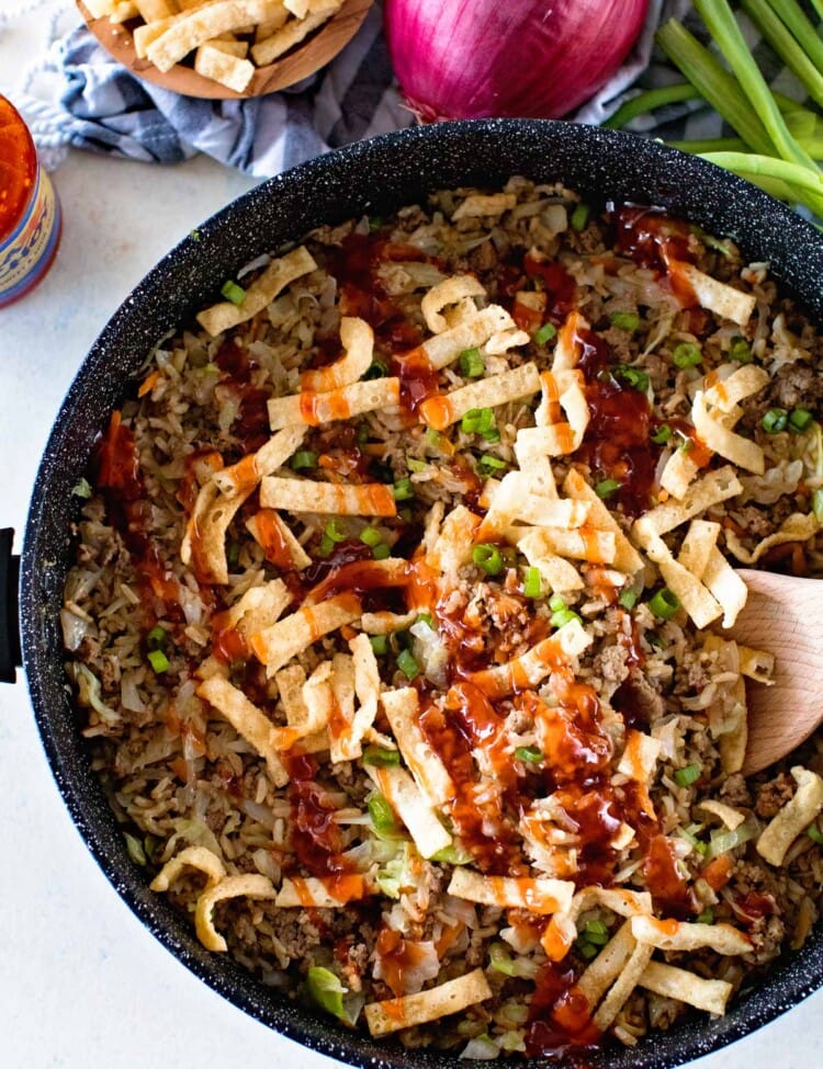 Overhead image of one pan egg roll in a bowl being stirred with a wood spoon next to a bottle of sauce, a bowl of crunchy strips, an onion, and some green onions