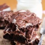 Three fudgy marshmallow brownies stacked on brown paper next to a glass of milk and a fork with brown text across the bottom reading fudgy marshmallow brownies