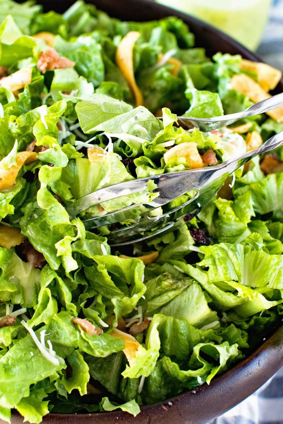 Silver serving tongs scooping up lettuce frito salad that's in a brown wood serving bowl.